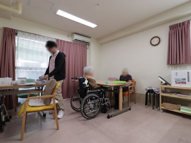 居室での日常風景 リビング ゆったりとした居室で、お客様が過ごされています。スタッフが手厚いサポートを行っており、温かい雰囲気が伝わってきます。
