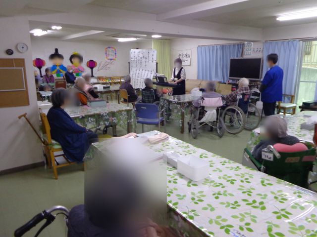利用者が集う多目的室 多目的室   テーブルを囲んで利用者の方が集まり、職員の方が前方で進行しています。テレビや掲示物もあり、交流や活動に使いやすい空間です。