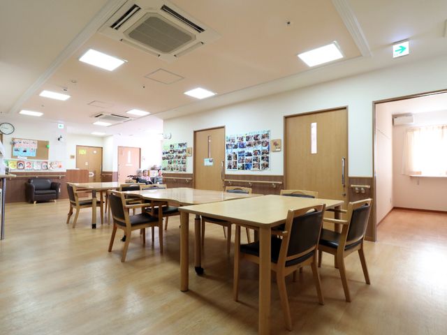 明るい共有スペース 食堂1 広々とした食堂は日当たりが良く、壁には楽しげな掲示物があります。テーブル周りには余裕があり、二人の利用者が対面で談笑しています。