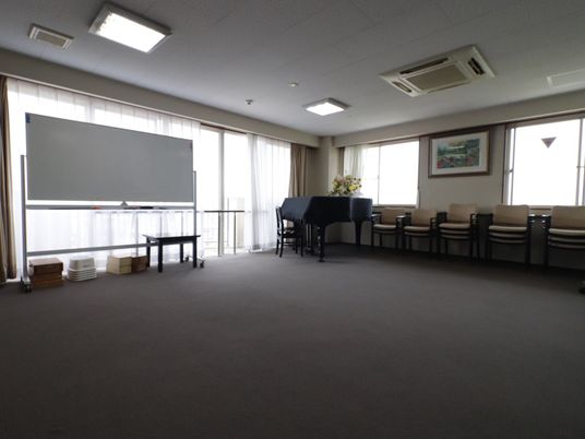 広々とした部屋には窓がある。窓際にはホワイトボードやピアノが置かれている。3つに重ねられた椅子が壁際に並べられている。