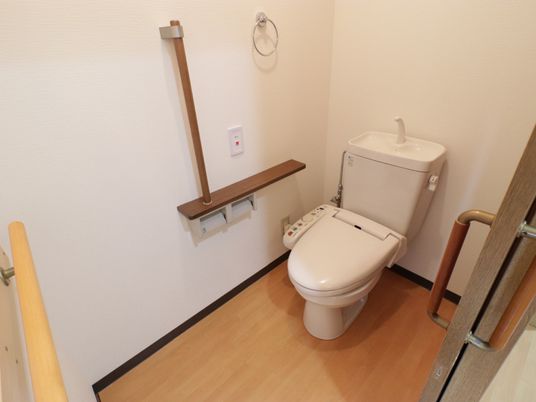 トイレには洋式便器が設置されている。壁側にはトイレットペーパーや手すり、呼び出しボタン、タオルハンガーが備え付けられている。