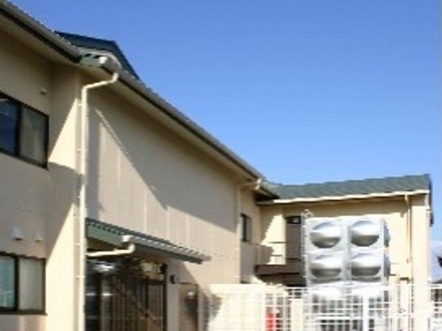 晴れた日の施設外観 玄関 陽光が照りつける広々とした駐車場を有する施設です。ゆとりある空間がお出迎えし、気持ちの良い職場環境を提供しています。