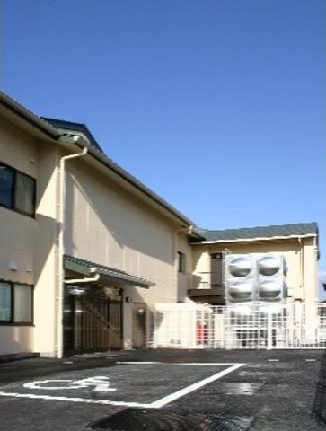 晴れた日の施設外観 玄関 陽光が照りつける広々とした駐車場を有する施設です。ゆとりある空間がお出迎えし、気持ちの良い職場環境を提供しています。