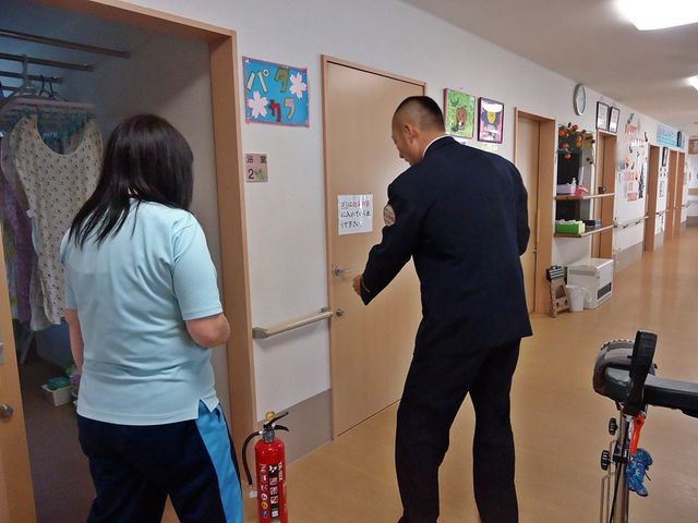 廊下で職員対応する様子 廊下   明るい廊下で、職員の方が扉を開けて確認しています。壁面の掲示物もあり、日常の動きが伝わる空間です。