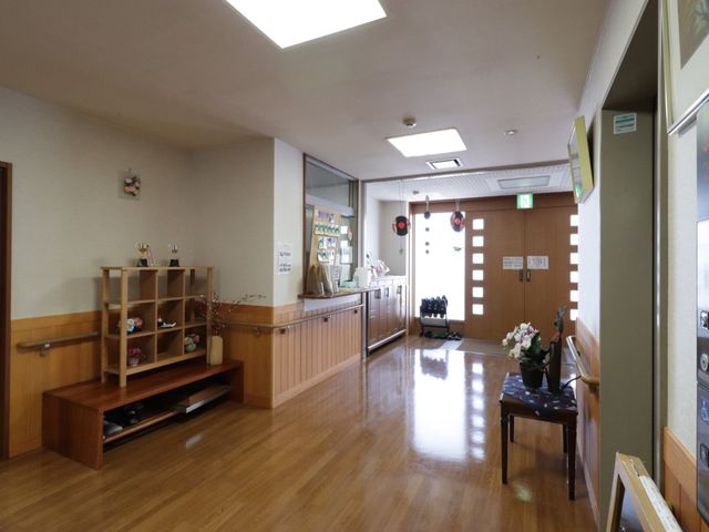 明るい廊下の風景 玄関 手すりと座席が備えられた廊下は、ご利用者様が安心して移動できる設計です。木目調の床が温かみのある雰囲気を演出しています。