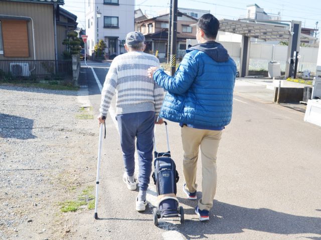 杖を使って歩く男性と若者の後ろ姿 外観 スタッフが親身になったサポートをしています。温かな雰囲気が伝わります。
