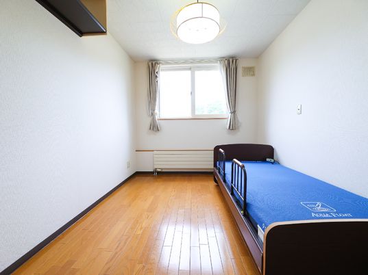 壁の一面にカーテンのついた窓がある青いマットレスが敷かれたベッドが置かれた白く四角い部屋