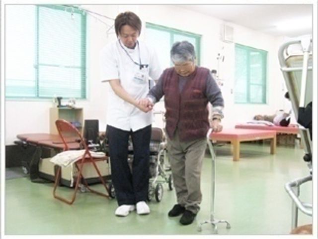 サポートを受ける様子の機能訓練室 機能訓練室 利用者がスタッフに支えられながら歩行訓練を行っています。丁寧なサポートで安心感があります。