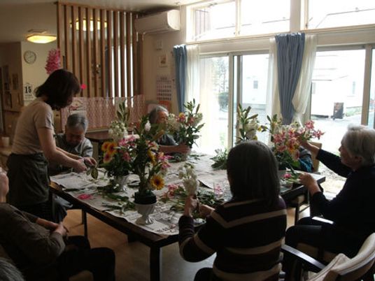 テーブルの上に新聞紙を広げ、フラワーアレンジメントを楽しんでいる高齢者たち。指導している女性もいる。