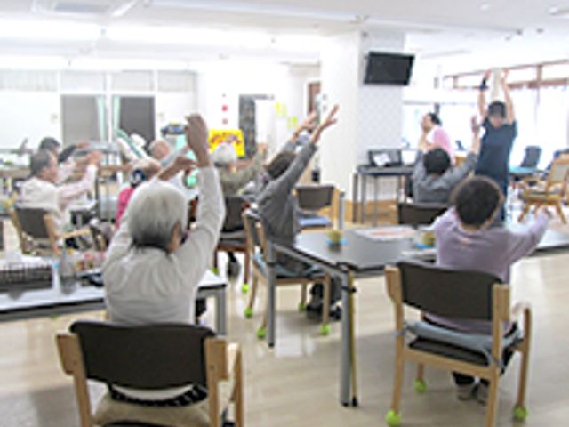 体操をする人々の集まる部屋 多目的室 自然光あふれる多目的室で、入居者の皆さんが体操を楽しんでおられます。リラックスした雰囲気です。