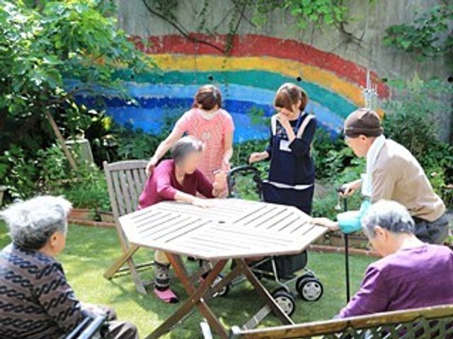 中庭での憩いのひととき 日常風景 緑あふれる中庭で、スタッフとの会話を楽しみながら、穏やかな時間を過ごされる方々の笑顔が印象的です。自然光の下、心地よい風を感じることができる空間です。