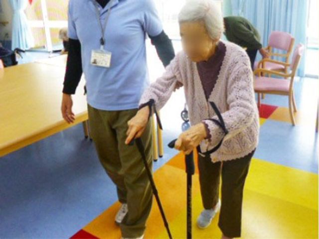 歩行訓練する高齢者と職員 リハビリ 職員の支援を受けながら、一歩一歩を大切に歩く高齢者の姿です。温かな雰囲気の中で、ご高齢者の健康維持をサポートしています。