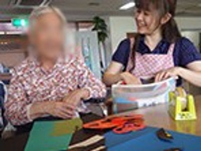 笑顔の職員と女性 ラウンジ 明るい共有スペースで、職員が利用者と一緒に作業を楽しむ様子です。温かい雰囲気が魅力的です。