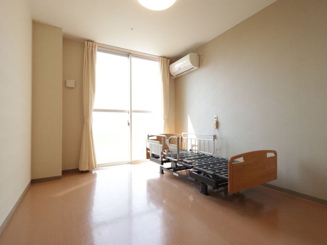 居室内のベッドと窓 居室  ゆったりとした室内のベッド横には大きな窓があり、充分な日差しが入る設計になっております。快適なプライベート空間でのびのびと過ごせる環境です。
