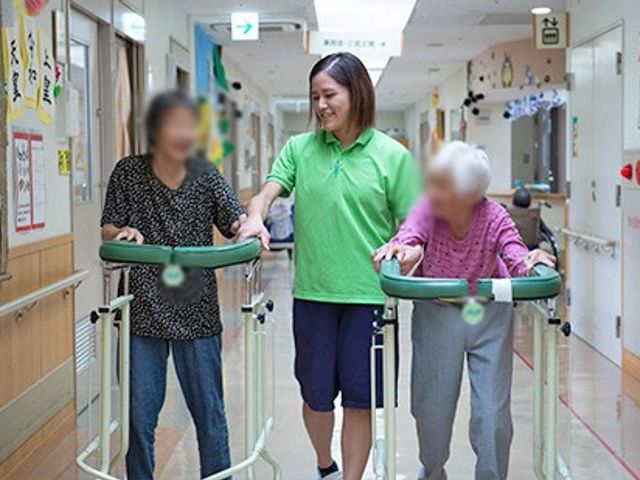 明るい廊下で人々が活動中 廊下 自然光が差し込む廊下で、スタッフが利用者をサポートしています。動きやすい工夫がされています。