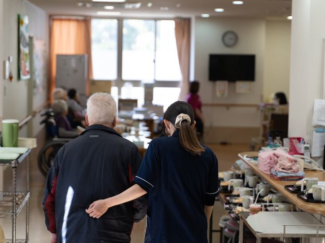 ケアを行うスタッフの後ろ姿 ダイニングルーム 職員が利用者様の手を取り、食事エリアへと伴走する光景から、温かい支援が感じられます。