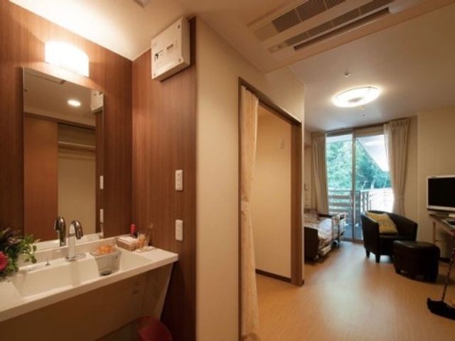 居室の入口と洗面台 居室 洗練された居室の一角には、実用的な洗面台と明るい窓が備わっており、清潔感が感じられます。窓の外には緑が見え、穏やかな時間をお過ごしいただけるでしょう。