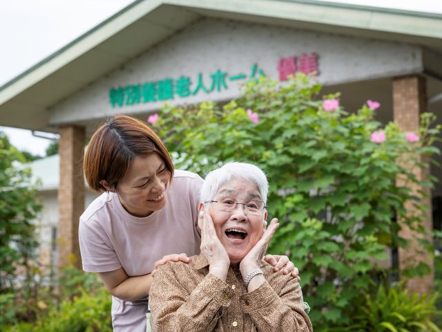 笑顔のシニアとスタッフの前庭 外観 花に囲まれた場所で、笑顔の利用者と親しみやすいスタッフが心温まるひとときを過ごしています。
