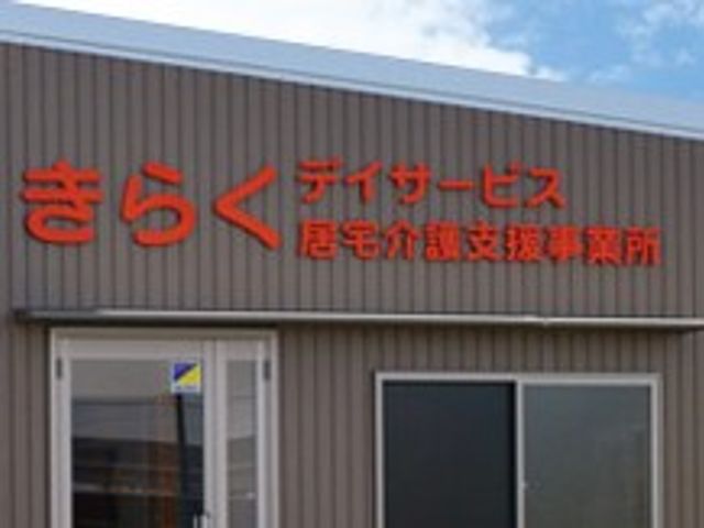 デイサービスの建物です 看板 外壁に赤い文字で施設名が掲示されています。建物はシンプルなデザインです。