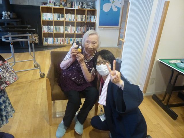 笑顔の高齢者とスタッフ 施設内の様子 手元にぬいぐるみを持ち、笑顔を見せる高齢の方と、ピースサインをするスタッフがいるアットホームな雰囲気のスペースです。この温かい環境でご一緒に業務をしませんか。