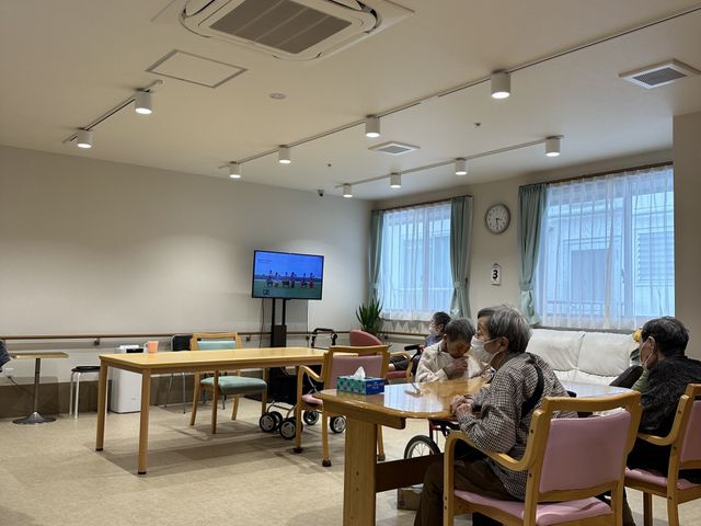 居室に集う人々 リビング 落ち着いた雰囲気の居室には、ゆったりと座れるソファや個々の椅子が配され、利用者の皆さんがくつろいでいます。木目調の内装が温かみを感じさせ、家庭的な安らぎの空間を提供しています。