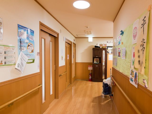 明るい廊下の様子 廊下 温かみのある木目調で統一された廊下は、ご利用者の安全と快適な移動をサポートしています。壁には掲示物があり、活動的な雰囲気が漂っています。