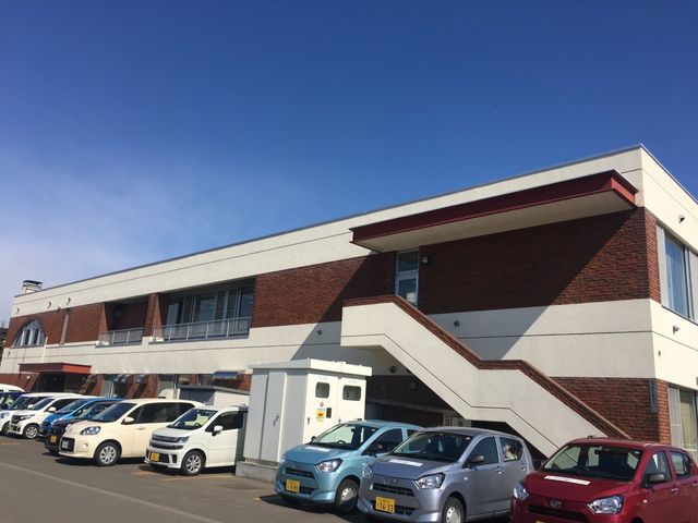 建物外観と駐車場 外観   青空の下に2階建ての建物が見え、前には車が並ぶ駐車場があります。出入口へ続く外階段も確認でき、来訪時の動線が分かりやすいです。