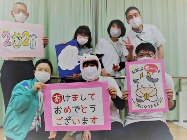 年始の集合場面、室内 多目的室   利用者さんとスタッフの皆さんが、年始の手作り掲示を持って並んでいます。明るい室内で、にぎやかな交流の様子が伝わります。