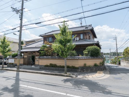 施設の写真 施設は道路に面して建てられている。歩行者通路には大きな木が植えられている。玄関入り口の横には施設の看板が取り付けられている。