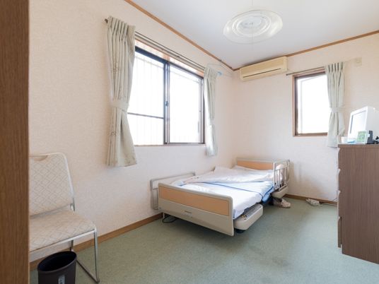 施設の写真 居室には窓が2つ設置されている。ベッドには手すりが取り付けられている。近くには背もたれ付きの椅子も置かれている。