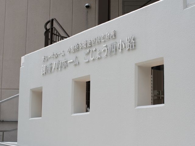 外壁に看板がある壁 ホームの看板 清潔感のあふれる白い外壁に目立つ看板があります。働く環境の第一印象として、明るく活気のある場所が伺えます。
