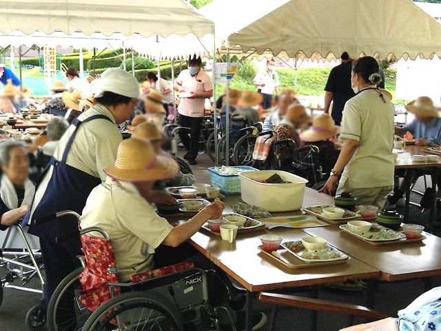 みんなで食事中の屋外 ダイニングルーム 爽やかな屋外で、居住者の方々が食事を楽しまれています。スタッフがサポートしています。