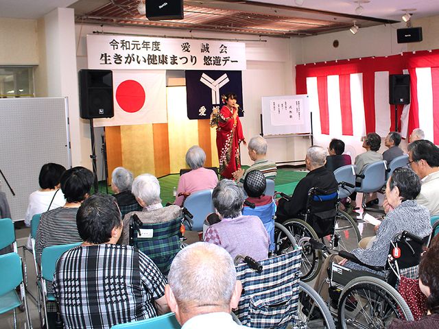 イベントを楽しむ利用者様 多目的室 椅子に座る方々が、前方のパフォーマンスに見入っている様子の共有スペースです。ゆったりとした時間をお過ごしいただけます。