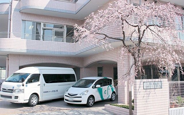 桜の下の車両と建物 外観   桜が迎える施設の正面。春を感じさせる優しい雰囲気のアプローチです。