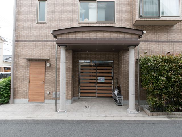 暖かみのあるエントランス 施設入り口 静かな住宅街に佇む、温かな雰囲気のエントランスです。清潔感があり、植栽も手入れされていて、安心して毎日を過ごせる空間が広がっています。
