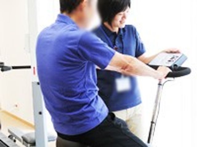機能訓練の様子 機能訓練室 利用者がスタッフとともに、トレーニング機器を使って体力作りを行う場面です。明るい環境です。