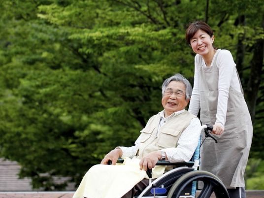 男性の入居者様が乗った車椅子に手を添える女性スタッフ。二人ともカメラに向かって笑顔を見せている。背後には緑豊かな木々が見える。