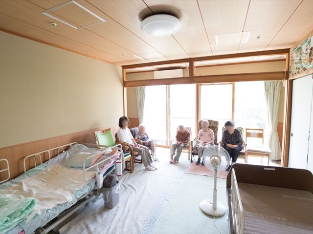 居室内の人々 日常 窓辺で穏やかに語らう人々がいる、明るく広々とした居室です。共有スペースでは、日々の憩いの時間を過ごされています。