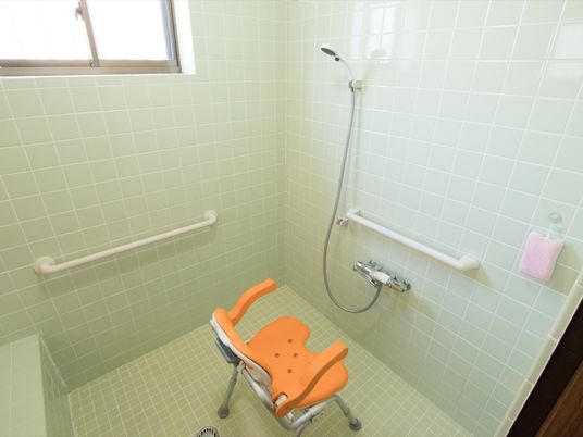 白を基調とした清潔感のある浴室である。洗い場にはひじ掛けと背もたれのついたオレンジ色の椅子が置かれている。