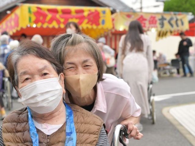 車いす利用の高齢者と介助者 スタッフイメージ   車いすの方に寄り添って笑顔を見せる高齢者の方々が写っています。あたたかな声かけや見守りの場面をイメージしやすいです。
