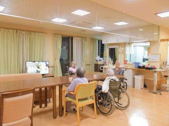 食堂で過ごす利用者 食堂   明るい食堂で、利用者の方がテレビを見ながらくつろいでいます。広めのテーブルがあり、車椅子でも過ごしやすそうです。