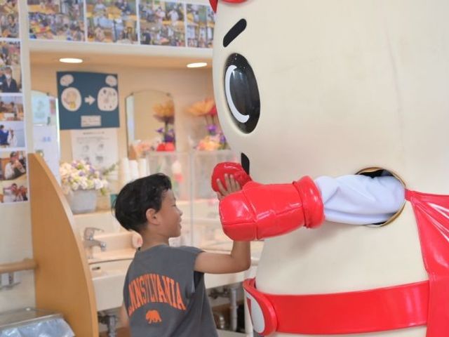 子どもが着ぐるみと触れ合うロビー ロビー   明るいロビーで、子どもが大きな着ぐるみと触れ合っています。温かい雰囲気の共有スペースがある職場をイメージしやすい場面です。