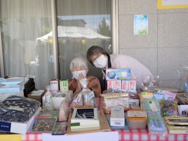 売店前のテーブル 外観   屋外の売店前に品物が並び、利用者と職員のやり取りが見られます。来訪者との交流を大切にする雰囲気が伝わります。