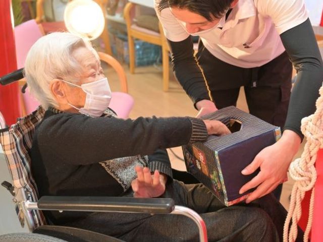 利用者支援の様子 ラウンジ   車椅子の利用者さんに職員さんが箱を手渡しており、会話しながら交流できる場面です。明るい共有スペースで、落ち着いた支援の様子が伝わります。