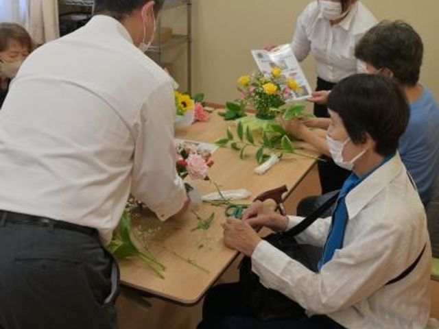 花を飾る交流室 多目的室   職員と利用者の方が机を囲み、花を扱いながら和やかに過ごしています。作業や交流の場として使いやすい、多目的な空間です。