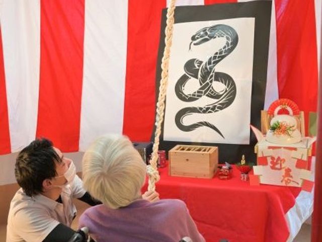 正月飾りのある多目的室 多目的室   紅白の装飾と干支の飾りが目を引く多目的室です。車椅子の方に寄り添う職員の様子もあり、季節の催しを楽しめる場面が伝わります。