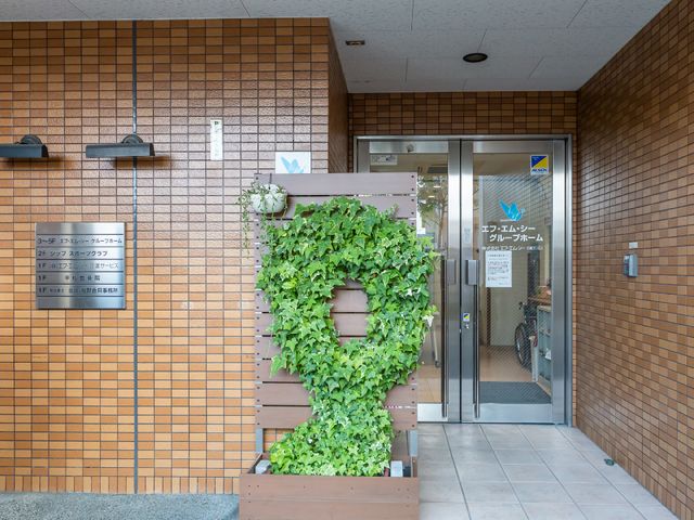 緑あふれるエントランス 玄関 清潔で開放的な入り口は、来訪者を暖かく迎え入れます。訪れる人々に安らぎを提供する落ち着いた空間です。