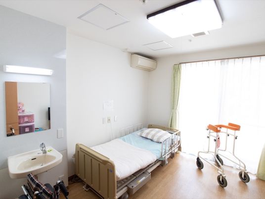 洗面台やエアコンが完備された居室である。介護用ベッドがあり、入居者様がご利用の歩行器や車椅子が置かれている。