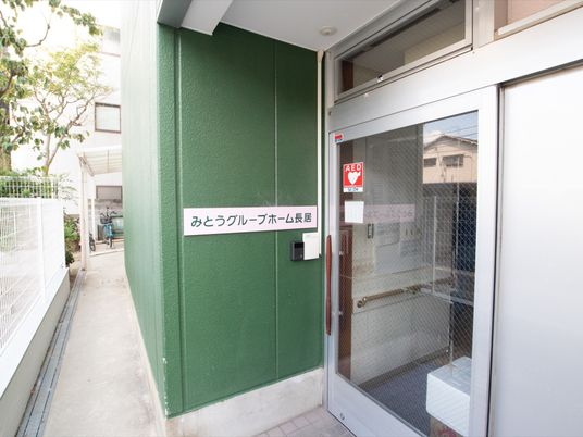 施設入口はグリーンの壁で、ガラス扉の引き戸が採用されている。インターホンや施設名称のプレートが設置されている。