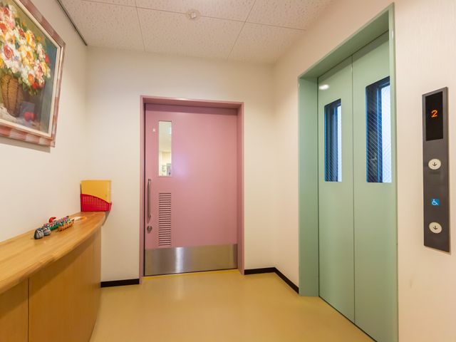 ピンクのドアと緑のエレベーター エレベーター ピンクのドアが印象的な明るい廊下です。周囲は清潔感があり、安心して過ごせる工夫が感じられます。